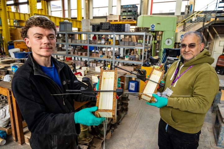 Two people hold up small steel devices for a photo taken inside a workshop